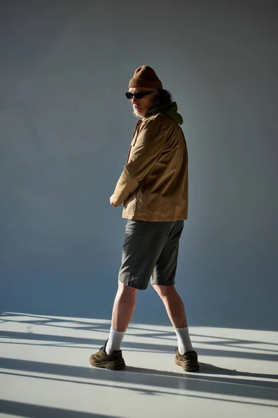 Volle Länge älterer Mann mit Hut, dunkler Sonnenbrille, Jacke und Shorts auf grauem Hintergrund mit Beleuchtung, hipster Stil älteres männliches Modell, modisches Alterskonzept — Stockfoto