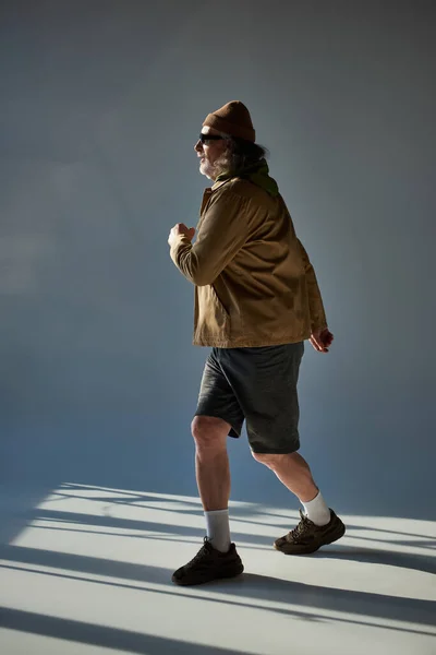 Side view of bearded and hipster style man in fashionable casual clothes walking on grey background with lighting, beanie hat, dark sunglasses, jacket and shorts, fashion and age concept — Stock Photo