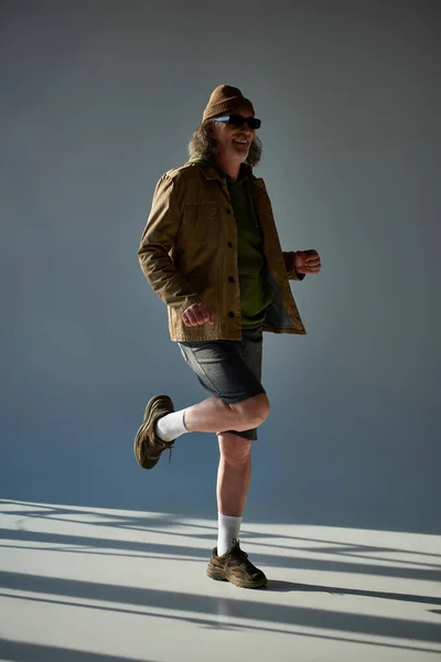 Full length of smiling and stylish senior male model posing on one leg on grey background with lighting, hipster fashion, dark sunglasses, jacket and shorts, happy and fashionable aging concept — Stock Photo