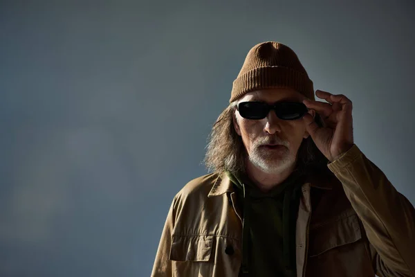 Modisch und hipster gestylt Senior mit Hut und brauner Jacke, dunkle Sonnenbrille und Blick in die Kamera auf grauem Hintergrund, alterndes Lebensstil-Konzept — Stockfoto