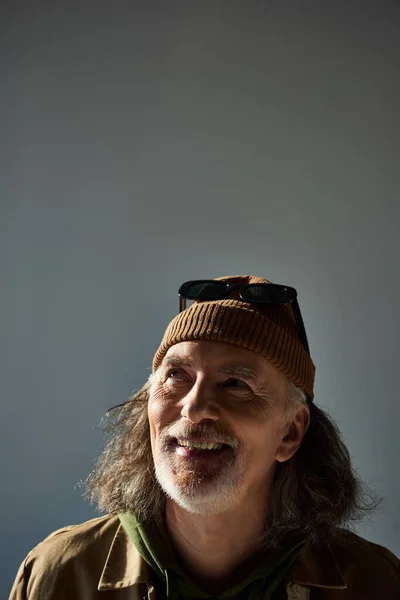 Portrait of cheerful and carefree senior man with grey hair and beard, wearing dark sunglasses on beanie hat, looking up on grey background, with copy space, happy and fashionable aging concept — Stock Photo