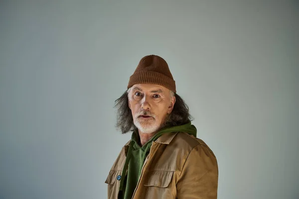 Thoughtful and discouraged hipster style senior man in beanie hat and brown jacket looking at camera while standing on grey background, aging population lifestyle concept — Stock Photo