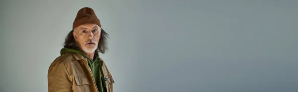 Вдумчивый пожилой человек в хипстерской одежде, шапочка и коричневая куртка, смотрящий на камеру на сером фоне, стареющая концепция образа жизни населения, баннер с копировальным пространством — стоковое фото