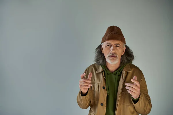 Upset and worried senior bearded man in beanie hat and brown jacket gesturing and looking at camera on grey background, hipster clothes, aging population lifestyle concept — Stock Photo