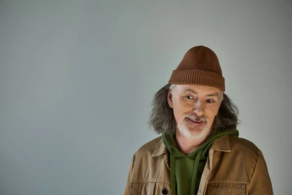 Hombre mayor de pelo gris y barbudo, reflexivo y sonriente, en gorro sombrero y chaqueta marrón sobre fondo gris, moda hipster, concepto de envejecimiento feliz y de moda - foto de stock