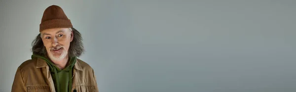 Homem senor pensativo e positivo sorrindo em fundo cinza, cabelos grisalhos, barba, chapéu de gorro, jaqueta marrom, estilo hipster, conceito de envelhecimento feliz e elegante, banner com espaço de cópia — Fotografia de Stock