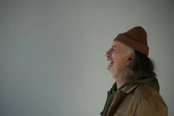 Vista lateral do homem idoso animado, cabelos grisalhos e barbudo em chapéu de gorro e casaco marrom rindo em fundo cinza, conceito de envelhecimento feliz e elegante, espaço de cópia — Fotografia de Stock
