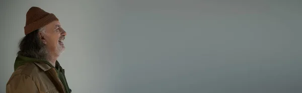 Profil des überglücklichen Seniors in braunem Sakko und Mütze lachend auf grauem Hintergrund, Hipster-Mode, fröhliches und modisches Alterskonzept, Banner mit Kopierraum — Stockfoto