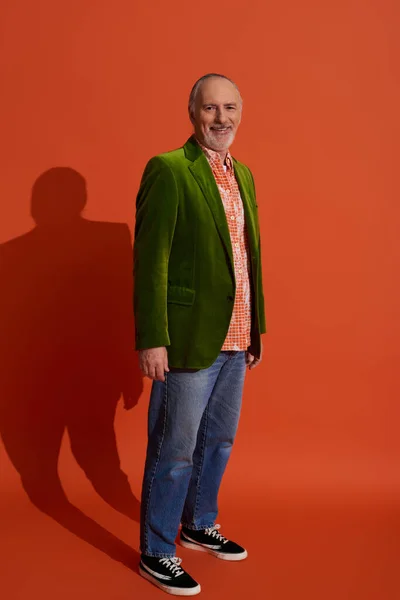 Full length of optimistic senior male model standing on red orange background and smiling at camera, trendy shirt, green velour blazer, blue denim jeans, personal casual style, happy aging concept — Stock Photo