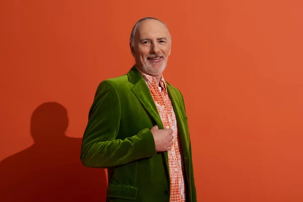 Hombre de pelo gris senior, carismático y alegre, posando en camisa de moda y chaqueta de terciopelo verde sobre fondo rojo anaranjado, mirando a la cámara, sonriente, concepto de envejecimiento positivo y de moda - foto de stock
