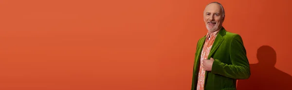 Hombre alegre, de pelo gris y barbudo mirando a la cámara mientras posando en chaqueta de terciopelo verde y camisa de moda sobre fondo rojo anaranjado, envejecimiento feliz, estilo anciano, moda y concepto de edad, bandera — Stock Photo