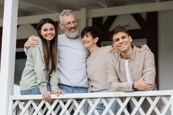 Fête des parents heureux, parents d'âge moyen embrassant la fille adolescente et le jeune fils adulte sur le porche de la maison d'été, célébration familiale, liaison, éducation parentale moderne, moments à retenir — Photo de stock