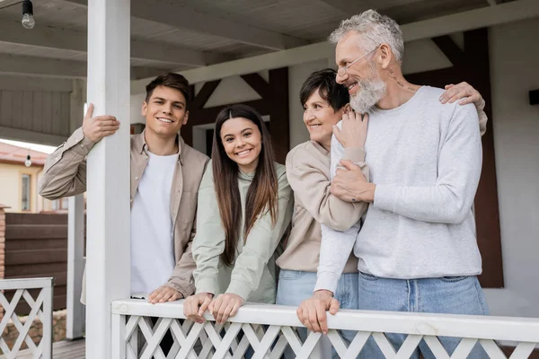 Parents day, middle aged parents hugging and looking away near teenage daughter and adult son on porch of summer house, family celebration, bonding, modern parenting, translation of tattoo: harm none — Stock Photo