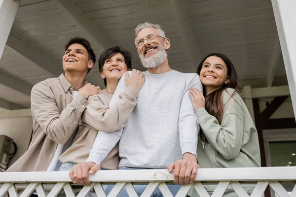 Celebration of parents day, middle aged parents hugging with happy daughter and son on porch of summer house, looking away, dreams, family reunion, bonding, modern parenting, moments to remember — Stock Photo