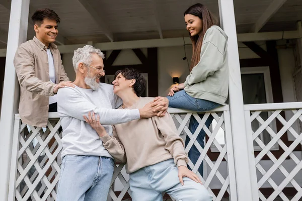 Parents day, joyful siblings looking at middle aged parents hugging and smiling on porch of summer house, family celebration, bonding, moments to remember, modern parenting, happy marriage — Stock Photo