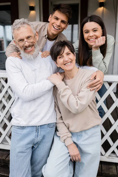 Family photo, happy parents day, joyful middle aged parents hugging near daughter and young adult son on porch of family house, celebration, bonding, moments to remember, modern parenting, June — Stock Photo