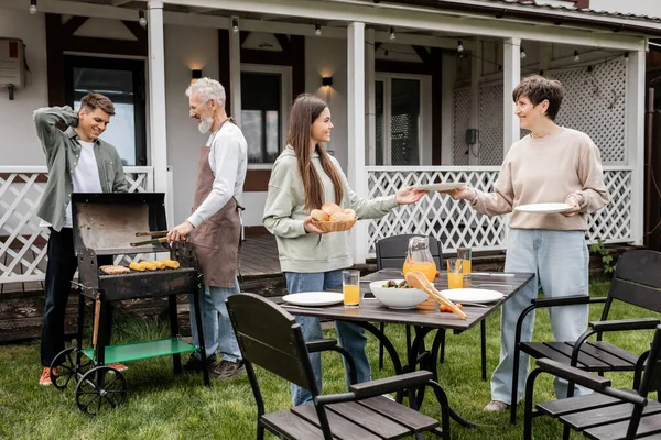 Parents day, cheerful teenage girl helping mother serving the table, father and son making bbq, grilling corn, backyard of family house, suburban life, family bbq party, celebration of summer — Stock Photo