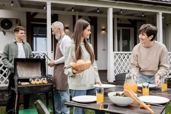 Parents day, happy teenage girl helping mother, holding buns, serving table, father and son making bbq, grilling corn, backyard of summer house, suburban life, family bbq party, celebration — Stock Photo