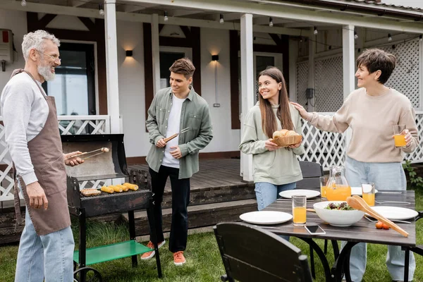 Parents day, happy teenage girl helping mother, holding buns, father and son making bbq, grilling corn, backyard of summer house, suburban life, family bbq party, bonding and celebration — Stock Photo