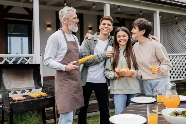 Family photo, parents day, happy middle aged parents having bbq party with teenage daughter and young adult son, father holding tongs with grilled corn, summer house, suburban life — Stock Photo