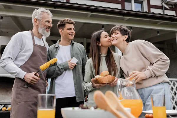 Parents day, happy middle aged parents, having family bbq party, teenage daughter kissing cheek of mother, siblings, father holding tongs with grilled corn, summer house, suburban life — Stock Photo
