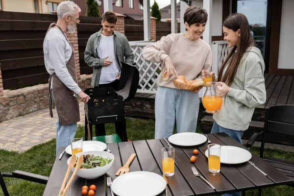 Celebration of parents` day, middle aged mother showing buns to teenage daughter with jug of orange juice, father and son preparing food on bbq grill, modern parenting, suburban life, summer — Stock Photo