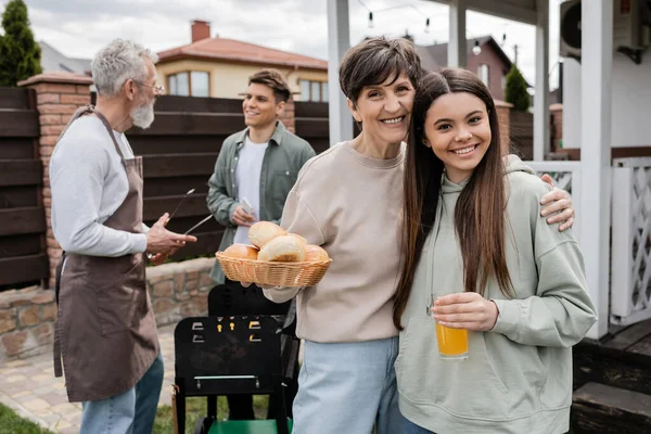 Modern parenting, celebration of parents day, happy middle aged mother holding buns and hugging with her teenage daughter, father and son preparing food on barbecue grill, backyard, modern parenting — Stock Photo