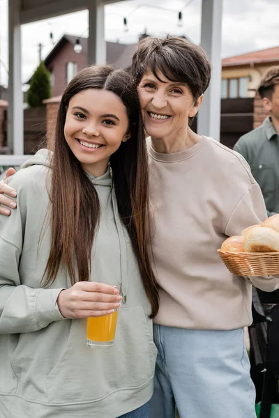 Modern parenting, celebration of parents day, happy middle aged mother holding buns and hugging cheerful teenage daughter, glass of orange juice, modern parenting, suburban life, summer — Stock Photo