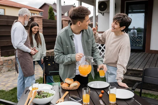 Celebration of parents day, modern parenting, happy middle aged mother talking to young adult son with jug of orange juice, father and daughter preparing food on bbq grill, summer, backyard — Stock Photo