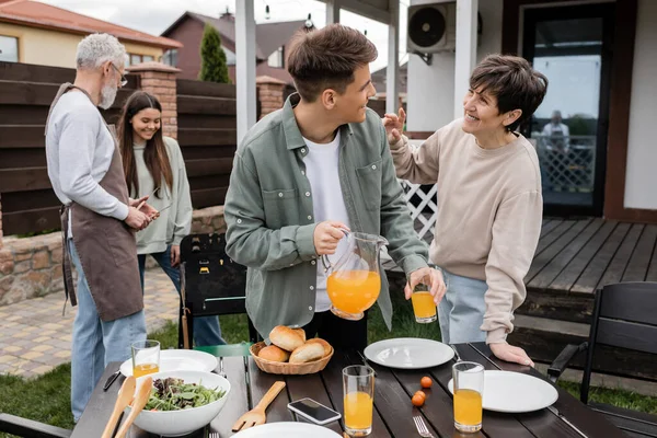 Celebración del día de los padres, crianza moderna, madre feliz de mediana edad hablando con el hijo adulto joven con jarra de jugo de naranja, padre e hija preparando la comida en la parrilla de barbacoa, verano, patio trasero — Stock Photo