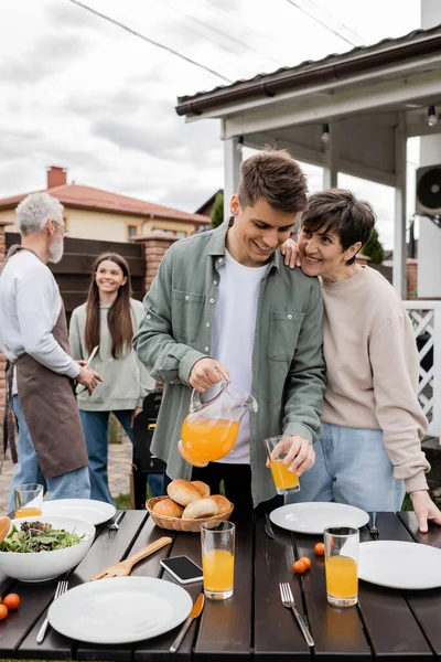 Celebration of parents day, modern parenting, happy middle aged mother leaning on young adult son pouring orange juice in glass, father and daughter preparing food on bbq grill, summer, backyard — Stock Photo