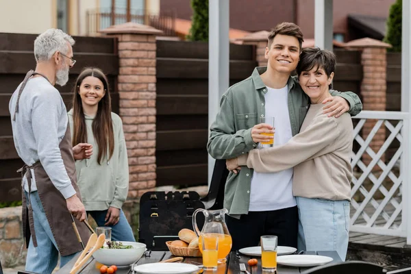 Happy parents day, modern parenting, cheerful middle aged mother hugging young adult son holding glass of orange juice, father and daughter preparing food on bbq grill, summer, backyard, celebration — Stock Photo