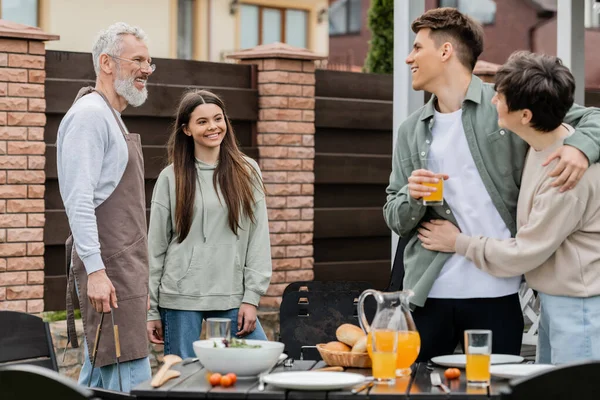 Happy parents day, modern parenting, cheerful middle aged mother hugging with young adult son, holding glass of orange juice, father and teenage daughter preparing food on bbq grill, summer — Stock Photo