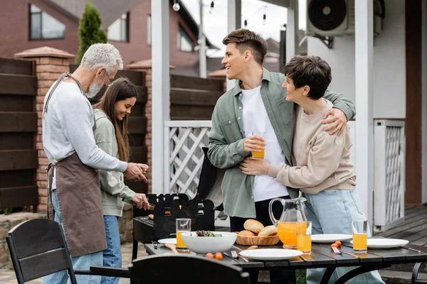 Modern parenting, happy parents day, middle aged mother hugging with cheerful young adult son holding glass of orange juice, father and teenage daughter preparing food on bbq grill, summer, backyard — Stock Photo