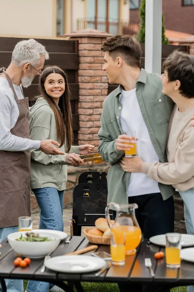 Modern parenting, happy parents day, father and teenage daughter preparing food on bbq grill, mother hugging with cheerful young adult son holding glass of orange juice, blurred foreground, backyard — Stock Photo