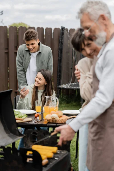 Happy teenage girl showing something on smartphone to young adult brother, digital age, father preparing food on bbq grill, barbecue party, parents day cerebration, backyard — Stock Photo