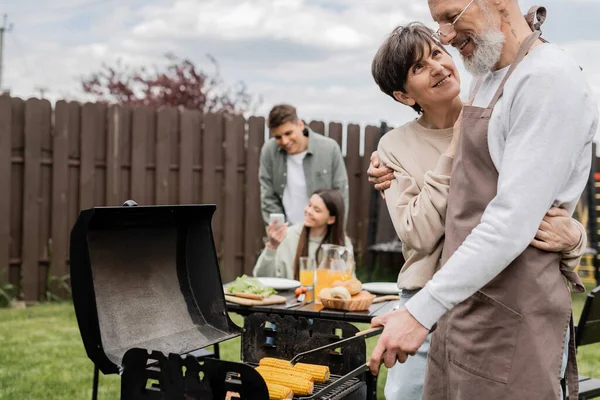 Parents day, middle aged couple hugging during family bbq party, bearded tattooed man holding tongs near bbq grill, grilling corn, daughter and son, translation of tattoo: harm none do what you will — Stock Photo