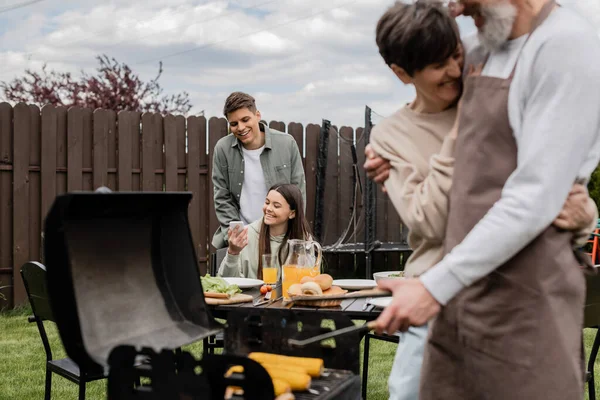Happy teenage girl showing something on smartphone to young adult brother, digital age, father preparing food on bbq grill, barbecue party, parents day cerebration, backyard, candid — Stock Photo