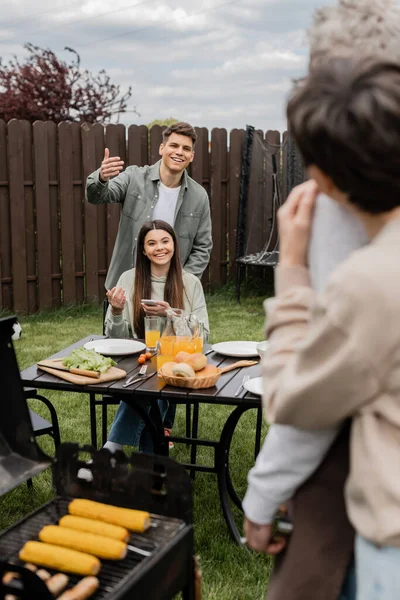 Parents day celebration, happy siblings looking at father and mother on blurred background, inviting to table, gesturing, family bbq, grill party, preparing food on grill bbq, modern parenting — Stock Photo