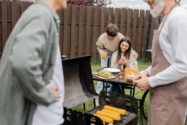 Cheerful teenage girl showing something on smartphone to young adult brother, digital age, father preparing food on bbq grill, barbecue party, parents day cerebration, backyard, candid — Stock Photo