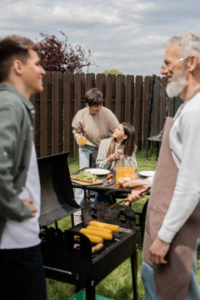Cheerful teenage girl holding smartphone and looking at mother digital age, father and son preparing food on bbq grill, barbecue party, parents day cerebration, backyard, blurred, candid — Stock Photo
