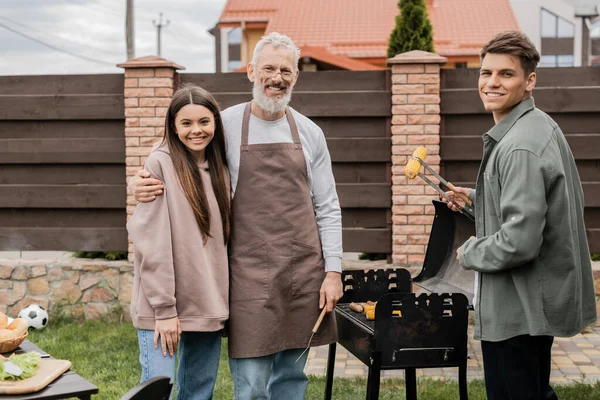 Happy parents day, middle aged father hugging teenage daughter, young adult son holding tongs and grilling corn, bbq party, family photo, looking at camera, backyard of summer house — Stock Photo