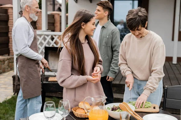 Parents day, candid, modern parenting, middle aged woman cutting lettuce near happy teenage daughter holding cherry tomatoes, making salad, father and son on blurred background, family bbq party — Stock Photo