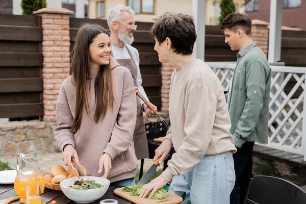 Cheerful mother and teenage daughter making salad, father and son preparing food on bbq grill, family house backyard, suburban life, happy parents day celebration, summer, june, candid — Stock Photo