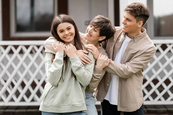 Happy parents day, cheerful mother hugging teenage daughter near young adult son on backyard of summer house, family celebration, bonding, moments to remember, modern parenting — Stock Photo