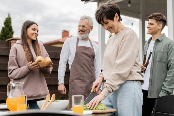 Positive middle aged woman cutting lettuce, teenage girl holding buns near father and brother during family grill party, bbq tongs, summer house, backyard, happy parents day concept — Stock Photo