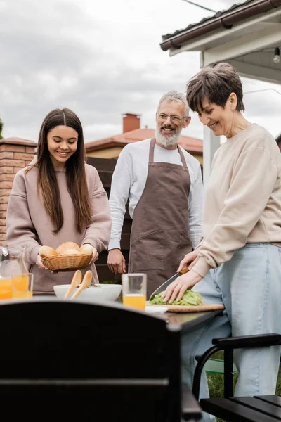 Joyful middle aged woman cutting lettuce, teenage girl holding buns near table and father in apron, grill party, bbq preparations, summer house, backyard, happy parents day concept — Stock Photo