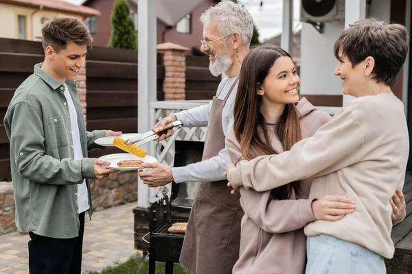 Happy mother and teenage daughter hugging on backyard of family house, father holding bbq tongs and putting grilled corn on plate of son, bbq party, parents day, translation of tattoo: harm none — Stock Photo