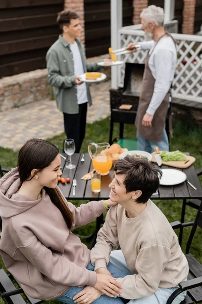 Joyful teenage girl talking to middle aged mother during family grill party, summer food, father and son preparing food on bbq grill, celebration of parents day concept, backyard of house — Stock Photo