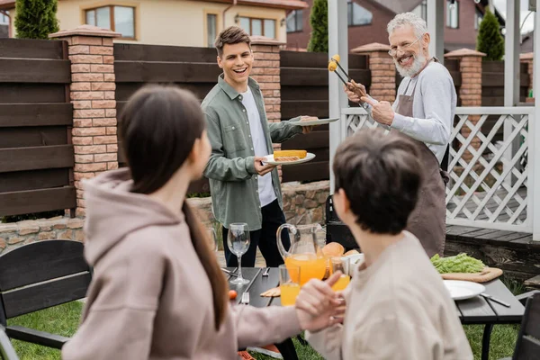 Bearded dad and his young adult son preparing food on bbq grill, family bbq party, grilled corn, middle aged man looking at wife and teenage daughter on blurred foreground, backyard — Stock Photo